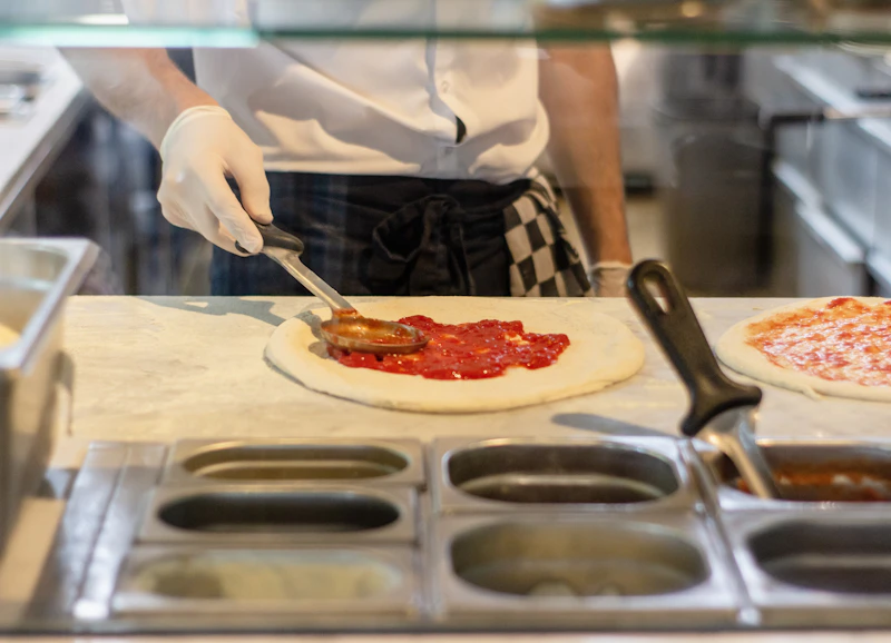 A chef spreading tomato sauce on pizza dough at a working kitchen counter.